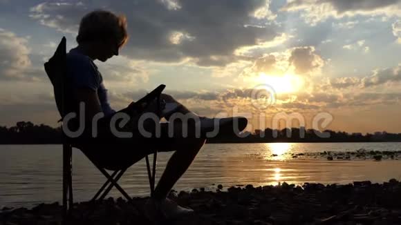 作家坐在美丽河岸日落的椅子上视频的预览图