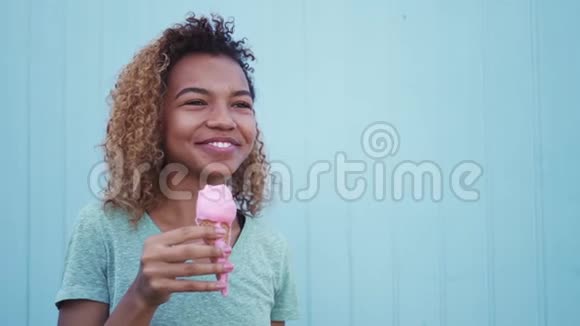 年轻的黑人女孩在蓝墙背景下吃粉色融化冰淇淋视频的预览图