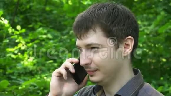 年轻人打电话用手机智能手机微笑户外帅哥在绿树上做特写镜头慢动作视频的预览图