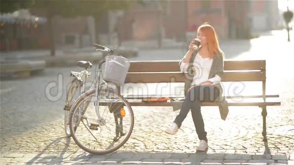 可爱的红头发女孩正在休息坐在城市的长凳上她推迟到一边喝咖啡视频的预览图