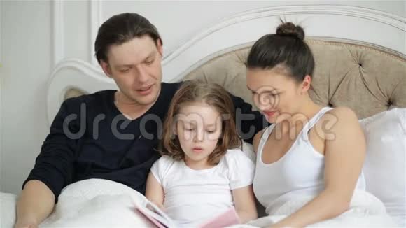 美丽的小女孩正在和她的父母一起读着躺在床上的书英俊的父亲正在亲吻他的聪明视频的预览图