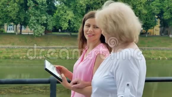 幸福的老年妇女和年轻妇女沿着海滨和平板电脑散步背景中的河流视频的预览图