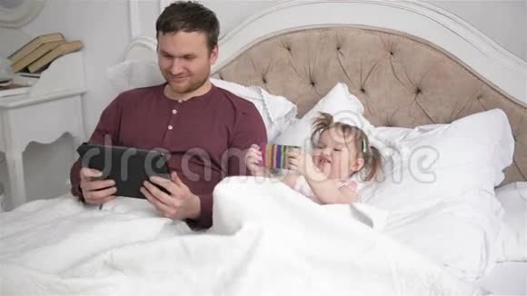 英俊的父亲和他的小女儿躺在床上使用电子设备小辫子的女孩子视频的预览图