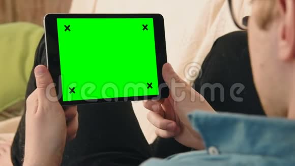 躺在沙发上看着绿色屏幕的数码平板电脑的年轻人视频的预览图