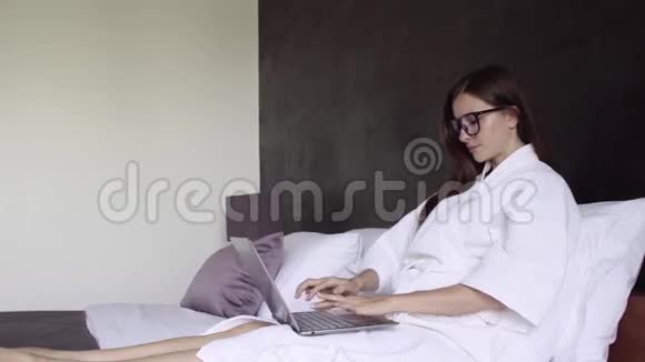 穿着白色浴袍和眼镜的女人在笔记本电脑上工作坐在床上女性微笑在电脑上工作商务视频的预览图