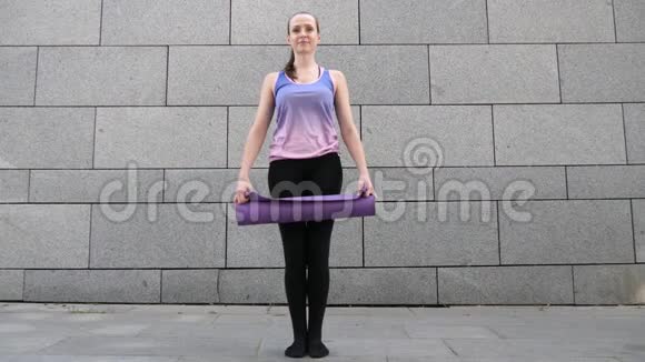 在灰色的城市背景下女人把瑜伽垫卷成相机自由健康和瑜伽理念近距离射击视频的预览图