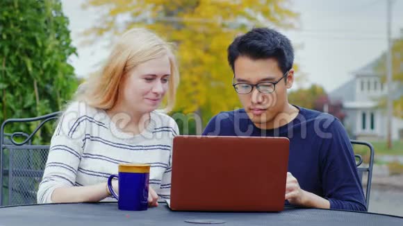 两个年轻的多民族朋友用笔记本电脑工作亚洲男人和高加索女人在夏季露台或咖啡馆视频的预览图