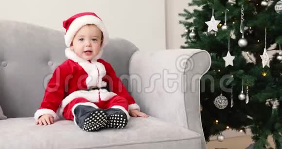 穿着圣诞老人套装的婴儿坐在沙发上和圣诞树视频的预览图