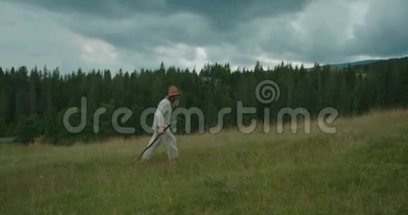 穿着旧衣服的老旅行者在恶劣的天气里拄着拐杖在美丽的青山上行走视频的预览图