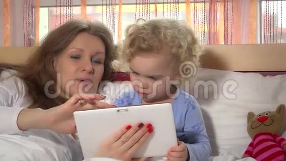 好奇的蹒跚学步的女孩和她妈妈在床上玩平板电脑视频的预览图