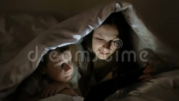 年轻女子妈妈和儿子一起在毯子下面的平板电脑上看有趣的电影视频的预览图