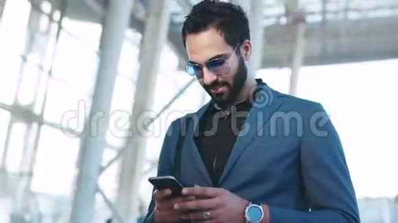 站在机场入口处的英俊留胡子的东方商人用他的电话和微笑接收消息视频的预览图