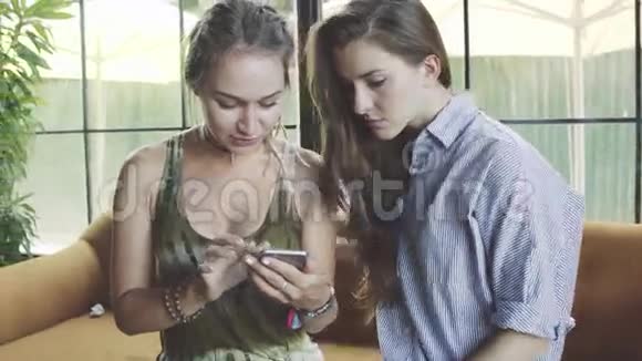 两个女朋友使用智能手机在舒适的咖啡馆里互相交谈视频的预览图