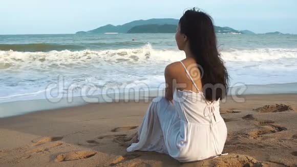 穿着白色连衣裙的亚洲女人坐在海边的沙滩上太阳沙子和波浪视频的预览图
