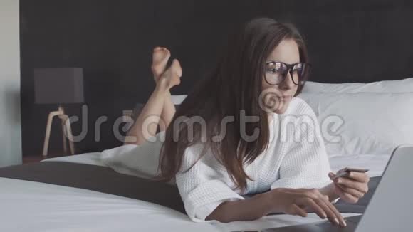 穿着白色浴袍和带信用卡和笔记本电脑的眼镜的女士躺在床上视频的预览图