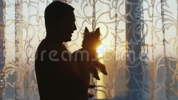 一个抱着狗的男人的剪影日落时分站在窗前视频的预览图