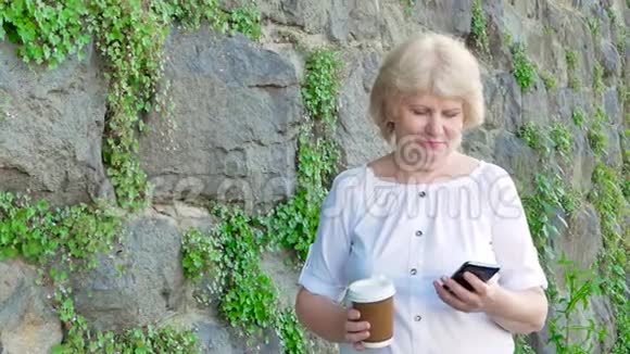 使用智能手机和喝咖啡的老年妇女背景中野生石头的复古墙视频的预览图