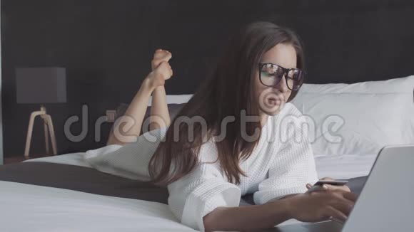穿着白色浴袍和带信用卡和笔记本电脑的眼镜的女士躺在床上网上购物视频的预览图
