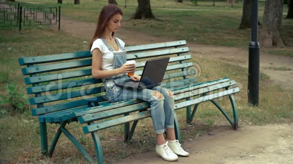 一个漂亮的女孩坐在公园里拿着笔记本电脑喝咖啡视频的预览图
