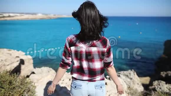 一位头发随风飘动的年轻女子举起双手站在美丽的海面上的悬崖上视频的预览图