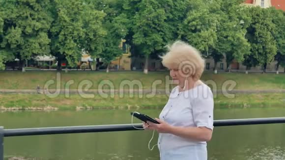 老年妇女沿着海滨散步使用带有耳机的平板电脑视频的预览图