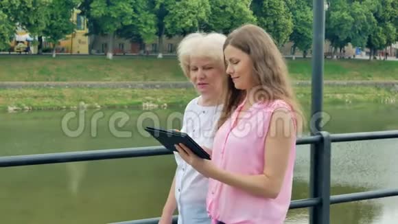 幸福的老年妇女和年轻妇女沿着海滨和平板电脑散步河流的背景视频的预览图