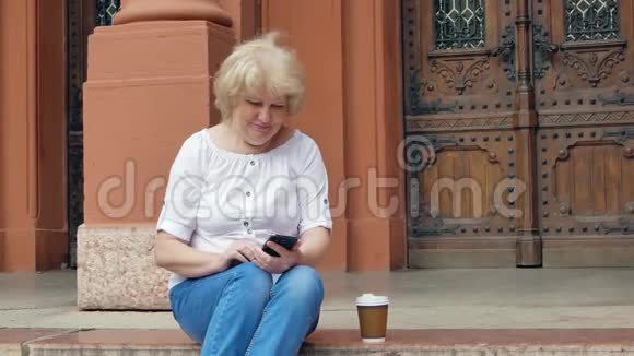 老妇人坐在台阶上用智能手机喝咖啡背景中的老式建筑视频的预览图