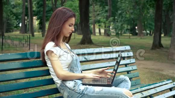 可爱的年轻女孩在公园里有一台笔记本电脑坐在长凳上视频的预览图