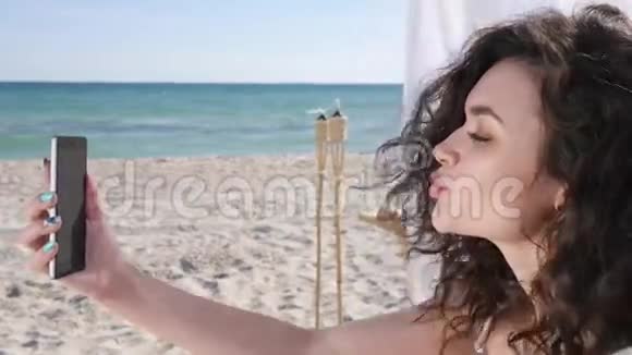 微笑的女孩在背景海洋和沙滩上自拍在海滩上自拍的年轻女子手机假日视频的预览图