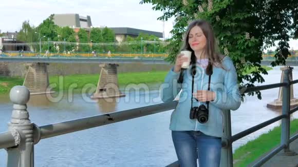 美丽的年轻女孩摄影师带着专业的相机在海滨河边和河边的栏杆旁喝咖啡视频的预览图