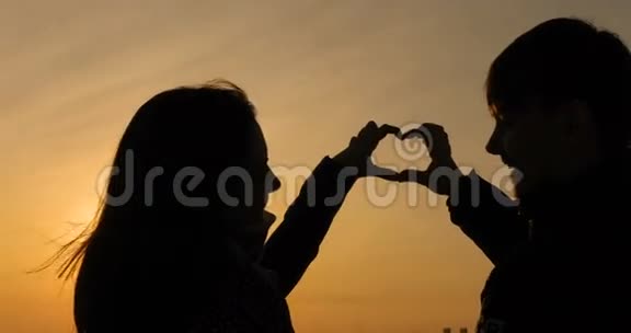 手里拿着太阳美丽的女孩和她的男朋友用他们的手在地平线上美丽的夕阳下塑造了心的形状视频的预览图