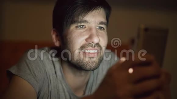 年轻的微笑男子使用智能手机浏览社交媒体晚上躺在家里的床上视频的预览图
