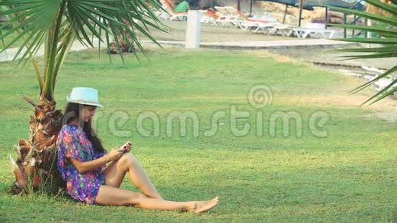 夏天一个皮肤黝黑的年轻女人长发坐在热带棕榈树下她用智能手机视频的预览图