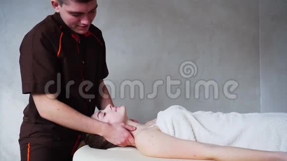 男按摩师用强壮的双手揉捏年轻女孩的脖子她躺在明亮的美容室的沙发上视频的预览图