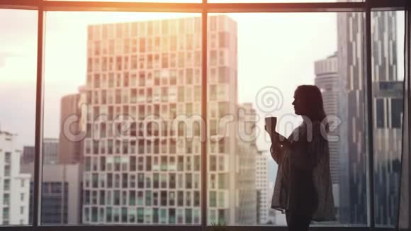 美丽的女人在窗边喝咖啡日落时有令人惊叹的城市景观慢动作3840x2160视频的预览图