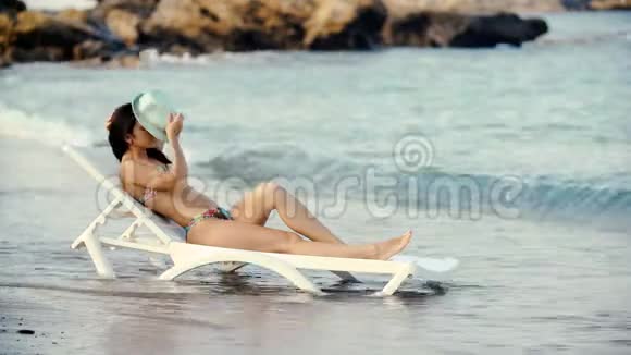 年轻漂亮的晒黑的女人长发披着比基尼躺在沙滩躺椅上视频的预览图