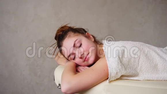 美丽的女人在按摩后放松低头看着躺在毛巾下的沙发上在明亮的医疗办公室视频的预览图