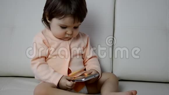 可爱的蹒跚学步的男孩坐在客厅的沙发上玩智能手机儿童学习如何使用视频的预览图