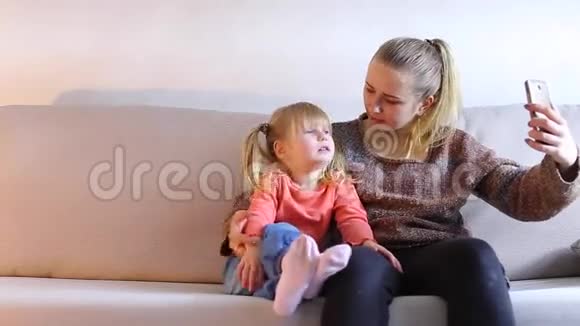 年轻的母亲和女儿坐在沙发上在电话上摆姿势自拍视频的预览图