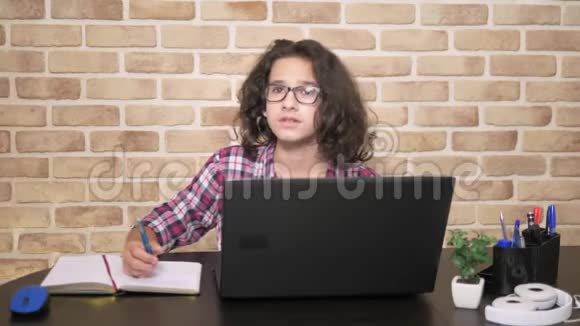 一个年轻漂亮的男孩头发卷曲黑发在笔记本电脑上工作在桌旁写字视频的预览图