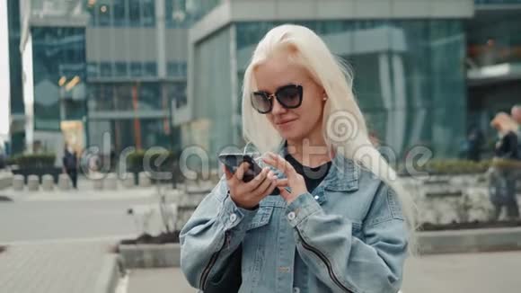 美丽的女人在走在街头模特金发的时候正在用智能手机上的应用程序发送一条短信视频的预览图
