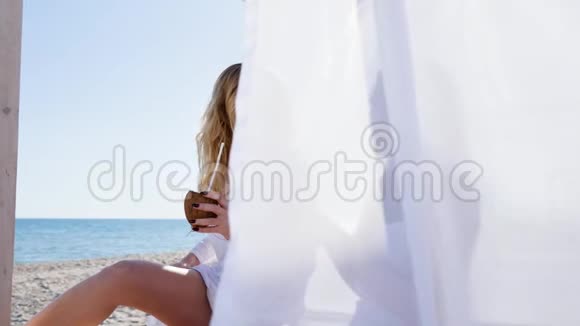 美丽的女孩在平房上夏天在海滩上休息风吹起白色的窗帘椰子在手里笑视频的预览图
