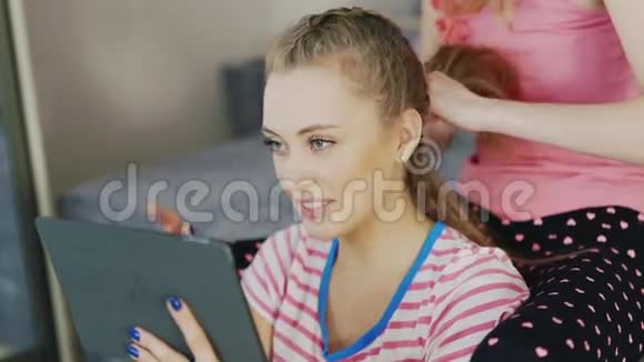 一个带平板电脑的年轻女人的肖像在家里放松女朋友做她的发辫视频的预览图