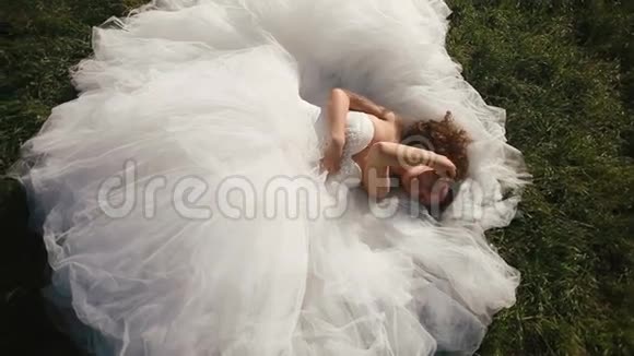 性感的年轻新娘穿着华丽的白色婚纱躺在绿草的草坪上诱人地看着镜头视频的预览图