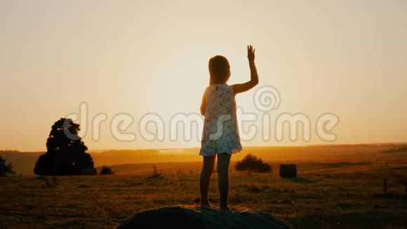 小女孩在日落的草地上挥手日落时分在田野里快乐的漂亮女孩视频的预览图
