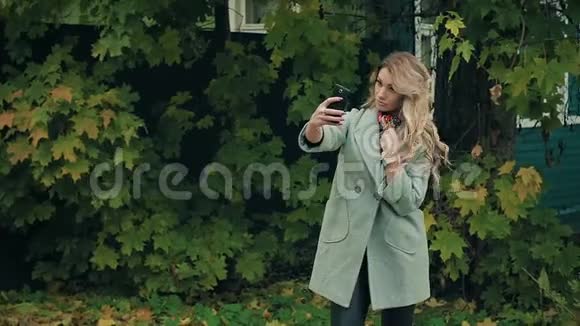 穿着灰色外套的金发美女在秋日公园的智能手机上自拍视频的预览图