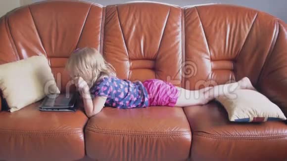 一个可爱的金发小女孩穿着所有粉红色的侧视图躺在沙发上在平板电脑上玩游戏4k视频的预览图