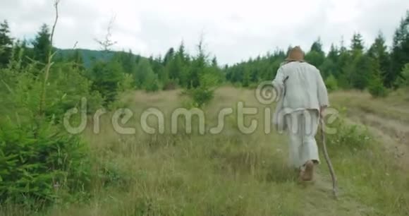 老人穿着旧衣服的后景是在拐杖的帮助下沿着山走的视频的预览图