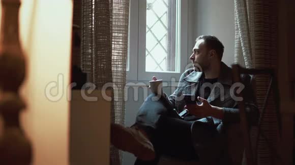 时髦的男人坐在扶手椅上看着窗户年轻男性在客厅里使用智能手机喝咖啡视频的预览图