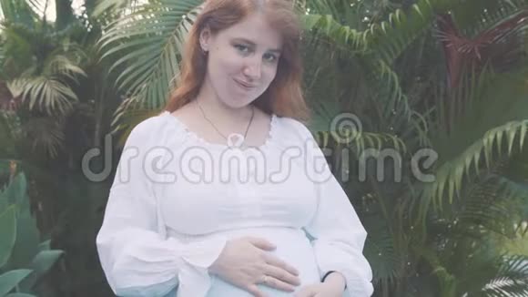 孕妇抚摸着她的小肚子在公园里的树间行走视频的预览图